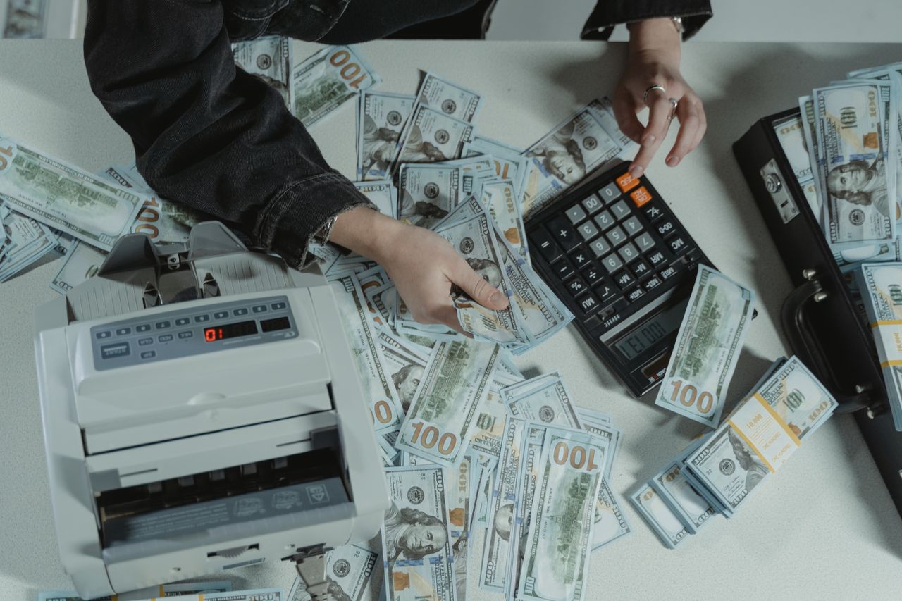 about-us-img Hands counting 100 dollar bills using a calculator and money counter on a table.