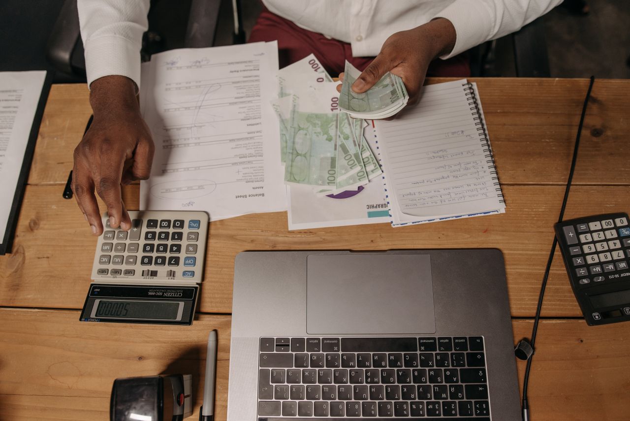 services-img-04 Close-up of a person counting cash with documents and a laptop in an office setting.